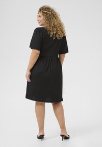 Czarna sukienka na krótki rękaw z ściąganym w pasie, teksturowanym materiałem, sięgająca do kolan, o zwiewnym kroju. Modelka w czarnych sandałach na obcasie z odkrytymi palcami.