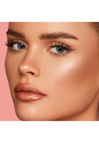 Primo piano di un volto con pelle liscia e abbronzata; sopracciglia definite; ciglia lunghe e scure; e labbra nude lucide su uno sfondo rosa.