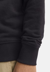 Schwarzer Baumwollpullover mit gerippten Bündchen und Saum sowie weicher Textur, auf einem Arm vor einem hellbeigen Hintergrund getragen.