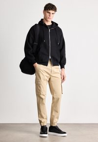 Zwarte rits hoodie, beige cargobroek met zijzakken en zwarte sneakers met witte zolen. Gladde textuur, comfortabele pasvorm en casual ontwerp.