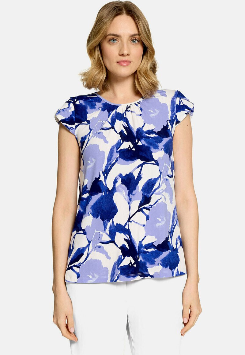 Femme portant un haut sans manches avec un imprimé floral abstrait bleu et blanc et un pantalon blanc sur un fond clair uni.