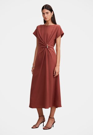 Femme portant une robe midi couleur rouille à manches courtes, une taille froncée avec un détail boucle et des sandales à talons noires à lanières.