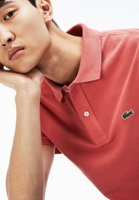 Camisa polo roja con una tela texturizada, cuello con solapa y tres botones. Presenta un logo de cocodrilo verde en el lado izquierdo del pecho.