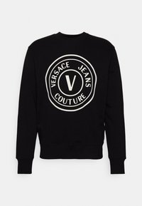 Schwarzer Pullover mit einem großen weißen Kreislogo, auf dem „VERSACE JEANS COUTURE“ und ein zentrales „V“-Design abgebildet sind. Mit gerippten Bündchen und Saum.