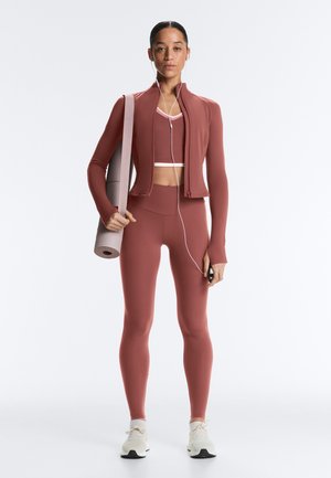 Giovane donna in abbigliamento sportivo color ruggine coordinato tiene un tappetino da yoga, indossa cuffie e tiene uno smartphone, in piedi su uno sfondo bianco uniforme.