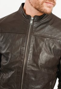 Brun läderjacka med hög krage, framtida dragkedja och slät textur. Har två sidofickor och enkla sömnadsdetaljer.