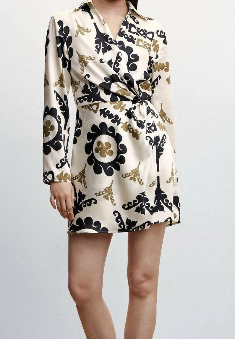 Femme portant une robe mini à manches longues couleur crème avec des motifs floraux abstraits noirs et dorés et un détail de nœud froncé à la taille.