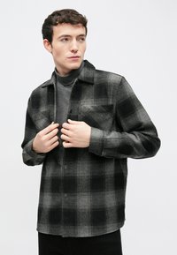 Μαύρο και γκρι φανελένιο πουκάμισο με καρό σχέδιο, δύο τσέπες στο στήθος, κλείσιμο με κουμπιά και γιακά, φορεμένο πάνω από γκρι ζιβάγκο.