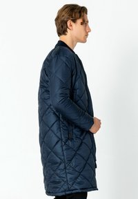 Marineblauer, gesteppter langer Mantel mit Reißverschluss, gerippten Manschetten und seitlichen Taschen. Verfügt über ein Rautenmuster und eine glatte Textur.