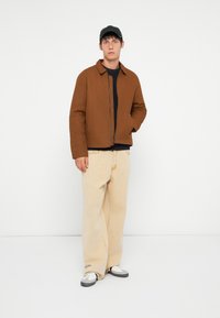 Brown zip-up jacket with a collar, navy sweater underneath, light beige wide-leg pants, and black and white sneakers. Soft fabric texture.
