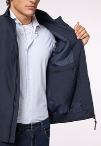 Giacca blu navy con chiusura a zip, tasca interna e texture morbida, indossata sopra una camicia a righe blu chiaro. Jeans in denim blu visibili.