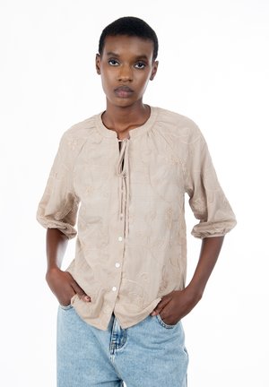 Junge Person mit kurzen Haaren, die ein beiges besticktes Hemd mit Knöpfen und hellblaue Jeans trägt, mit den Händen in den Taschen vor weißem Hintergrund steht.