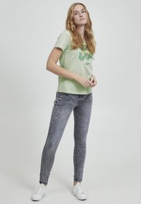 Camiseta de algodón verde claro con diseño de hojas, combinada con jeans skinny gris oscuro. El calzado son zapatillas blancas con detalles texturizados.