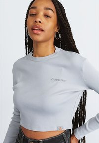 Jauna moteris su ilgomis nertinėmis kasomis, vilkinti šviesiai pilką trumpą, raukšlėtą viršų su "Dickies" logotipu ir auskarais žiedais, fono vientisame paveikslėlyje.