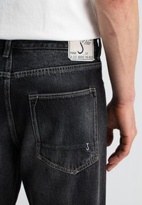 Rechte Hand und Hüfte einer Person, die schwarze Jeans trägt, mit einer Gesäßtasche und sichtbarem Markenlabel über dem Bund.