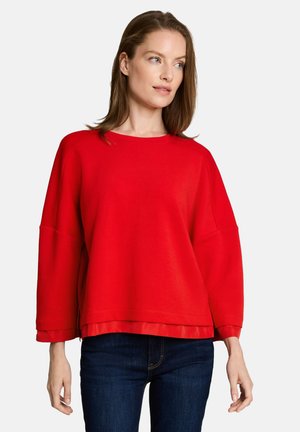 Woman with light skin and straight brown hair wears red long-sleeve loose top over dark blue jeans, looking slightly to her right.