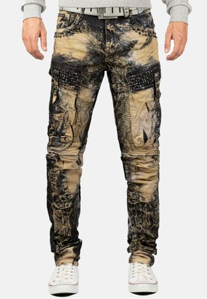 Herren Slim-Fit Cargo-Hose mit braun-schwarzem Batikmuster, mit Nieten besetzte Taschenklappen, Reißverschlussdetails, kombiniert mit weißen Sneakers und grauem Sweatshirt.