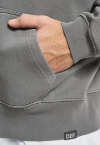 Sweat-shirt gris avec poche avant, tissu texturé, poignets et ourlet côtelés, avec une petite étiquette noire "DEF" en bas.