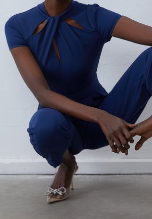 Kvinde i knæbøjning iført en marineblå jumpsuit med udskæringsdetalje og gennemsigtige spidse hæle med en rhinsten sløjfeaccessory.