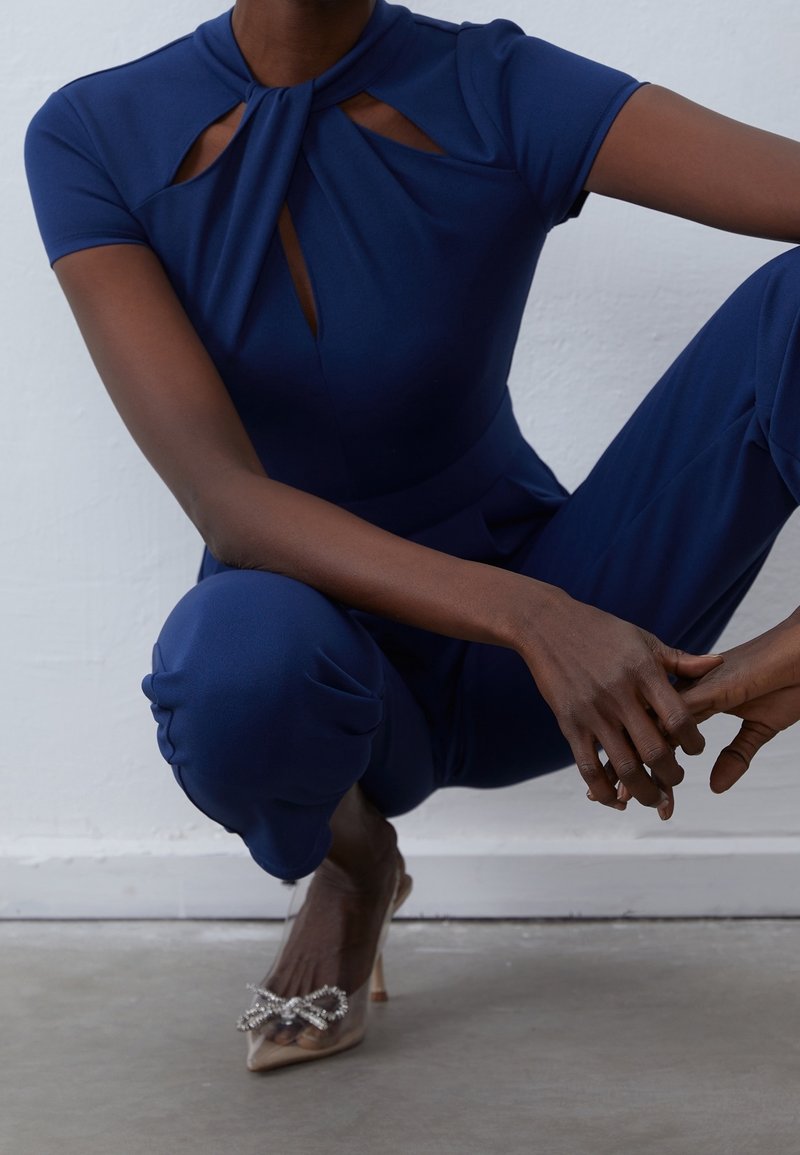 Femme accroupie portant une combinaison bleu marine avec des découpes et des talons transparents pointus ornés d'un nœud en strass.