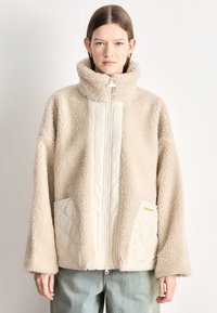 Beežist võltskarvaga jope kõrge krae, tõmbluku kinnituse ja kahe tikitud ees taskuga. Omadusteks on kuldne riistvara ja lõdvestunud lõige.