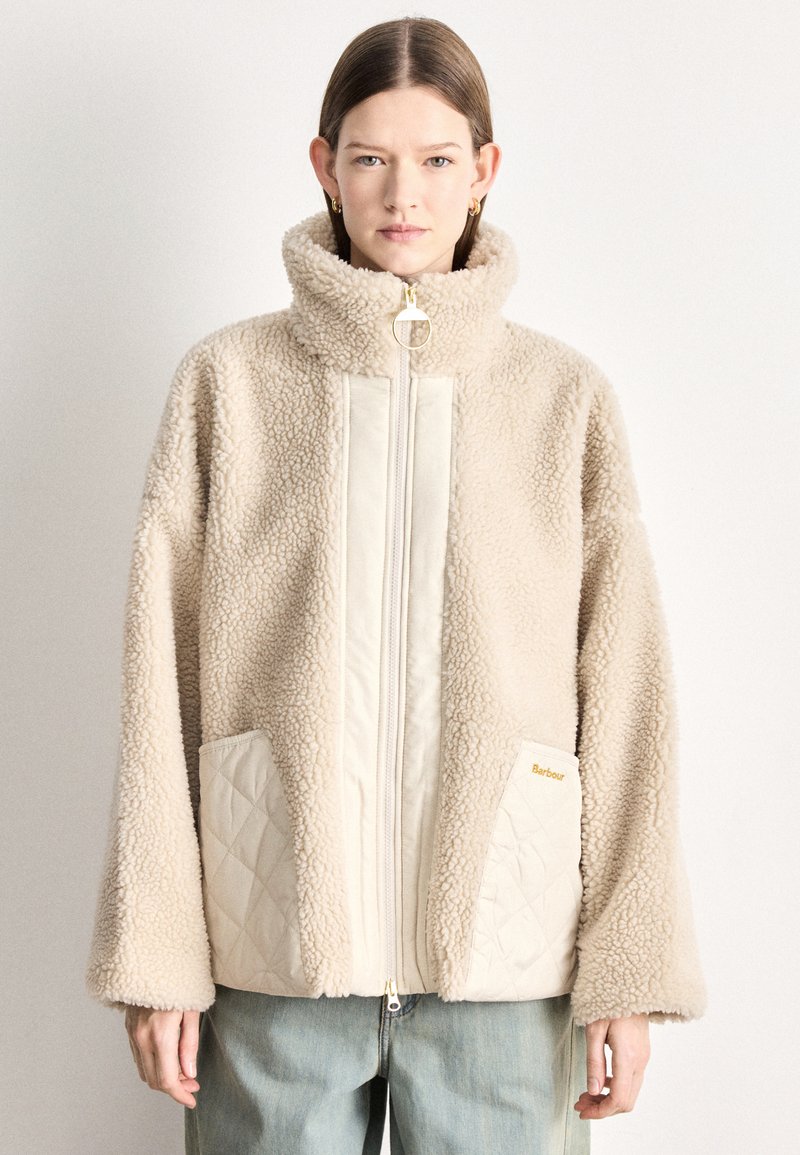 Beežist võltskarvaga jope kõrge krae, tõmbluku kinnituse ja kahe tikitud ees taskuga. Omadusteks on kuldne riistvara ja lõdvestunud lõige.