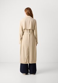 Beige Trenchcoat mit doppelreihigem Design, tailliertem Bund und Rückenschlitz; getragen über weit geschnittenen dunklen Jeans.