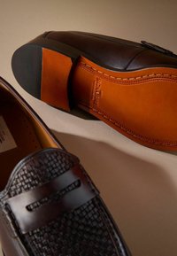 Braune Lederloafers mit gewebter Textur, mit einer glatten, runden Zehenform und einer kontrastierenden orangen Ledersohle. Geprägtes Logo auf der Innensohle.