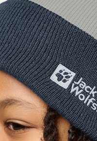 Beanie lavorato a maglia di colore blu scuro con fascia grigia chiara. Presenta una texture a coste e il logo ricamato di Jack Wolfskin con grafica della zampa.