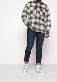 Camisa de cuadros en negro, blanco y beige, con dos bolsillos delanteros, combinada con jeans azul oscuro y zapatillas blancas. Corte relajado y estilo casual.