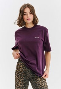 Fialové bavlněné tričko s krátkým rukávem a kulatým výstřihem, na hrudi s malou bílou grafikou řas.