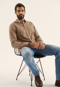 Chemise boutonnée à carreaux marron et orange, associée à un jean bleu clair et des chaussures en cuir marron, assis sur une chaise en fil métallique.