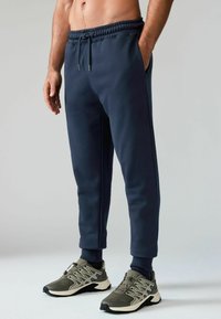 Marineblaue Jogginghose aus weichem Stoff mit elastischem Bund und Bündchen, ausgestattet mit seitlichen Taschen und einem Kordelzug. Kombiniert mit grauen Turnschuhen.