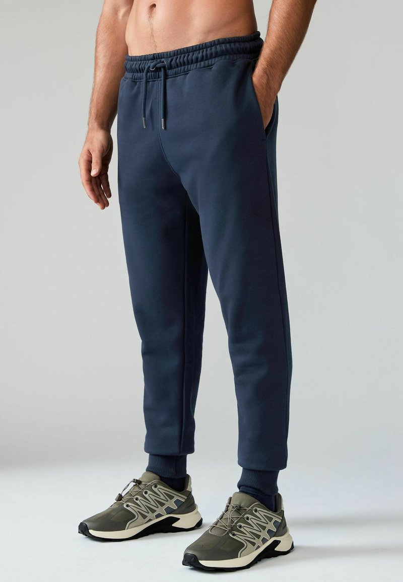 Marineblaue Jogginghose aus weichem Stoff mit elastischem Bund und Bündchen, ausgestattet mit seitlichen Taschen und einem Kordelzug. Kombiniert mit grauen Turnschuhen.
