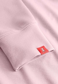 Lichtroze sweatshirtstof met een zachte textuur, voorzien van een omgeslagen manchet en een klein rood label met een grafisch ontwerp.