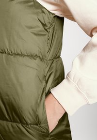 Olivově zelená prošívaná vesta s hladkou, lesklou texturou, která má boční kapsy a prošívaný design. Pod ní je světlá košile s dlouhými rukávy.