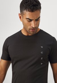 T-shirt preto de mangas curtas, feito de tecido suave, com um logo branco "PUMA" vertical do lado esquerdo e um design de gola redonda.