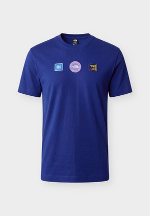 Mand med mørkt, mellemlangt hår iført en blå t-shirt med tre farverige logoer på brystet og sorte bukser, står med fronten mod kameraet.