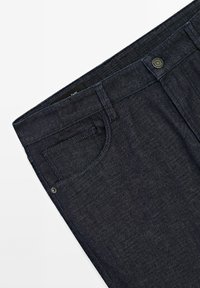 Tmavě modré džíny z denimu s plochým pasem, přední kapsou a kovovým zdobným knoflíkem. Hladká textura a mírně zúžený střih nohavic.