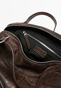 Brun lædertaske med en struktureret yderside, lynlås, og sort stofindvendig. Har en indvendig lynlåslomme og et lædermærke.