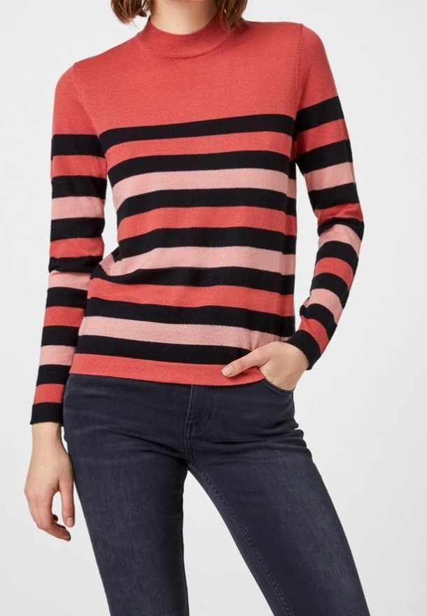 Femme portant un pull à manches longues avec des rayures horizontales noires, corail et roses, associée à un jean foncé ajusté, posant avec une main dans la poche.