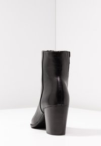 Botín negro de cuero con tacón cuadrado, presentado desde detrás sobre una superficie blanca y la esquina de una pared gris claro.