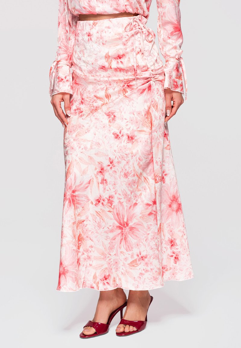 Femme portant une jupe midi fleurie rose et blanche et un haut à manches longues assorti avec des sandales rouges à talons ouverts.