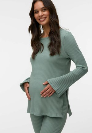 Uśmiechnięta kobieta w ciąży, ubrana w prążkowany, szałwiowozielony długi rękaw i dopasowane spodnie, delikatnie trzymająca brzuch obiema rękami.
