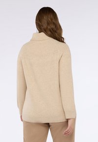 Beżowy sweter z dzianiny z ryflowanym kołnierzem, z teksturowanym tylnym karczkiem i długimi rękawami, w połączeniu z beżowymi spodniami. Miękki, lekki materiał.