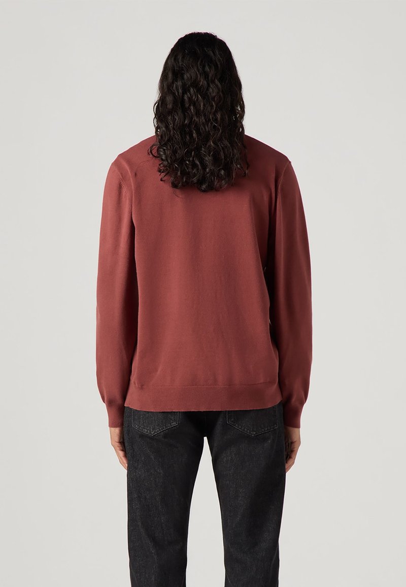 Sweatshirt de couleur rouille, à manches longues et encolure ronde. Tissu doux et poignets côtelés. Porté avec un jean foncé, affichant une coupe décontractée.