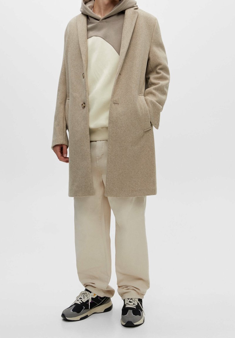 Cappotto beige in misto lana con bavero a rever, due bottoni, tasche frontali, indossato sopra una felpa grigia con cappuccio e pantaloni color crema, abbinato a sneaker nere.