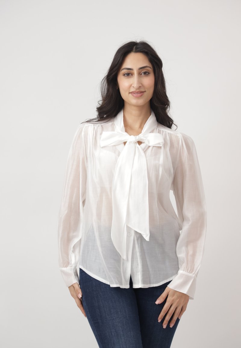 Frau mit langem dunkelbraunem Haar, die eine durchsichtige weiße Bluse mit großer Schleife und dunkelblaue Jeans trägt, steht vor hellem Hintergrund.