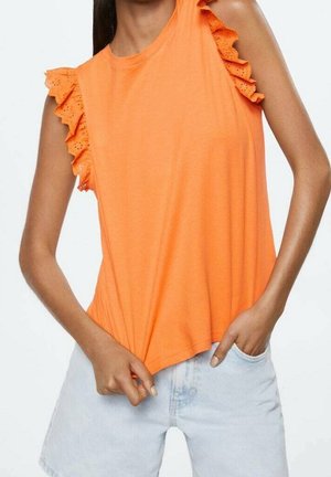 Mujer que lleva una parte superior sin mangas de color naranja brillante con volantes y adornos de encaje de ojales en los hombros y pantalones cortos de mezclilla azul clara.