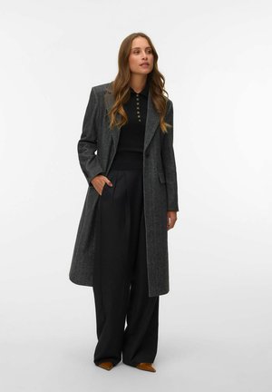 Femme portant un long manteau gris, une chemise noire boutonnée, un pantalon noir large et des chaussures brunes pointues, debout avec une main dans la poche.